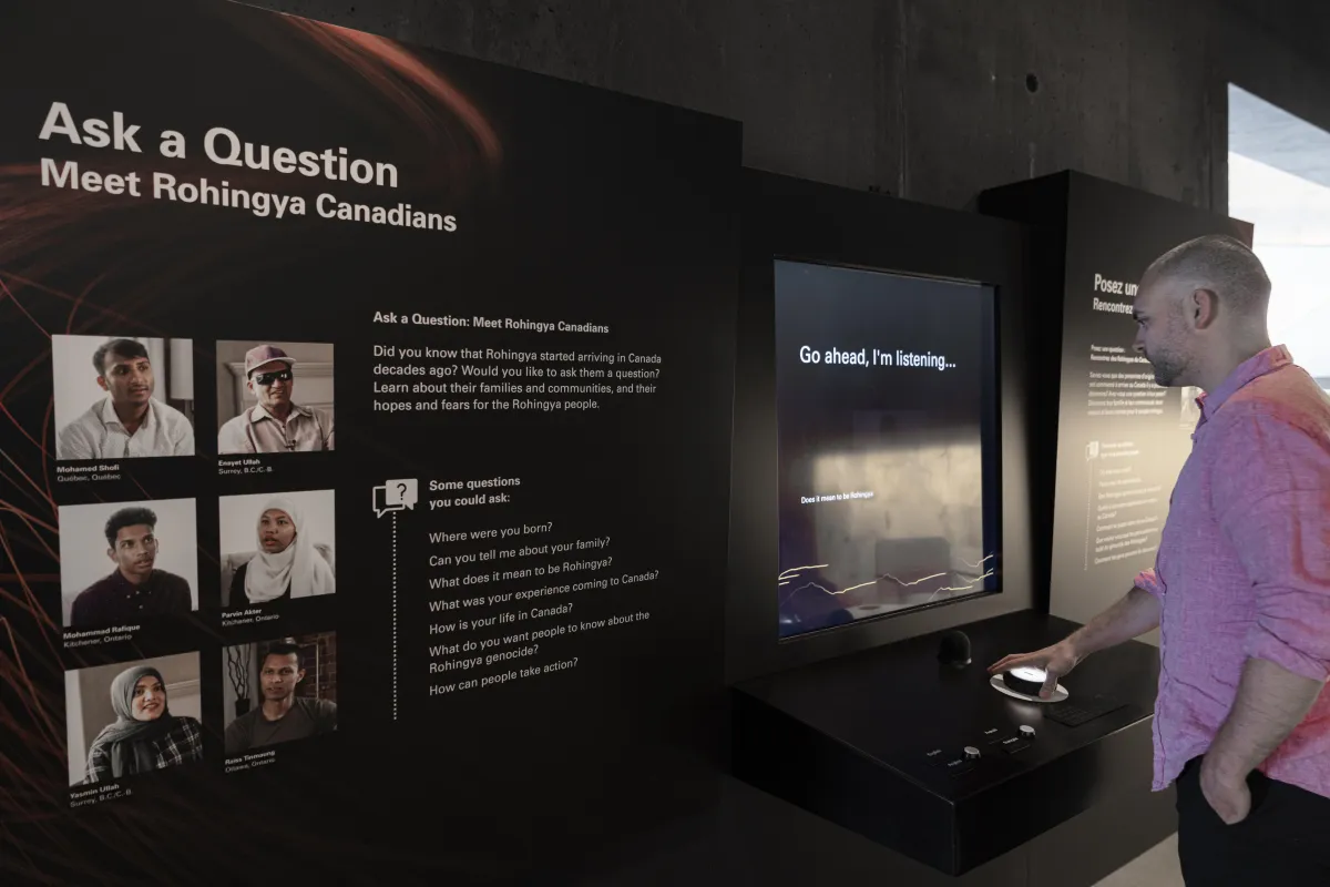 Un homme touche un gros bouton devant un écran vidéo qui affiche en anglais les mots : « Go ahead, I'm listening. » Au premier plan, il y a un panneau avec des photos de six personnes sous le titre, en anglais, « Ask a Question, Meet Rohingya Canadians ».