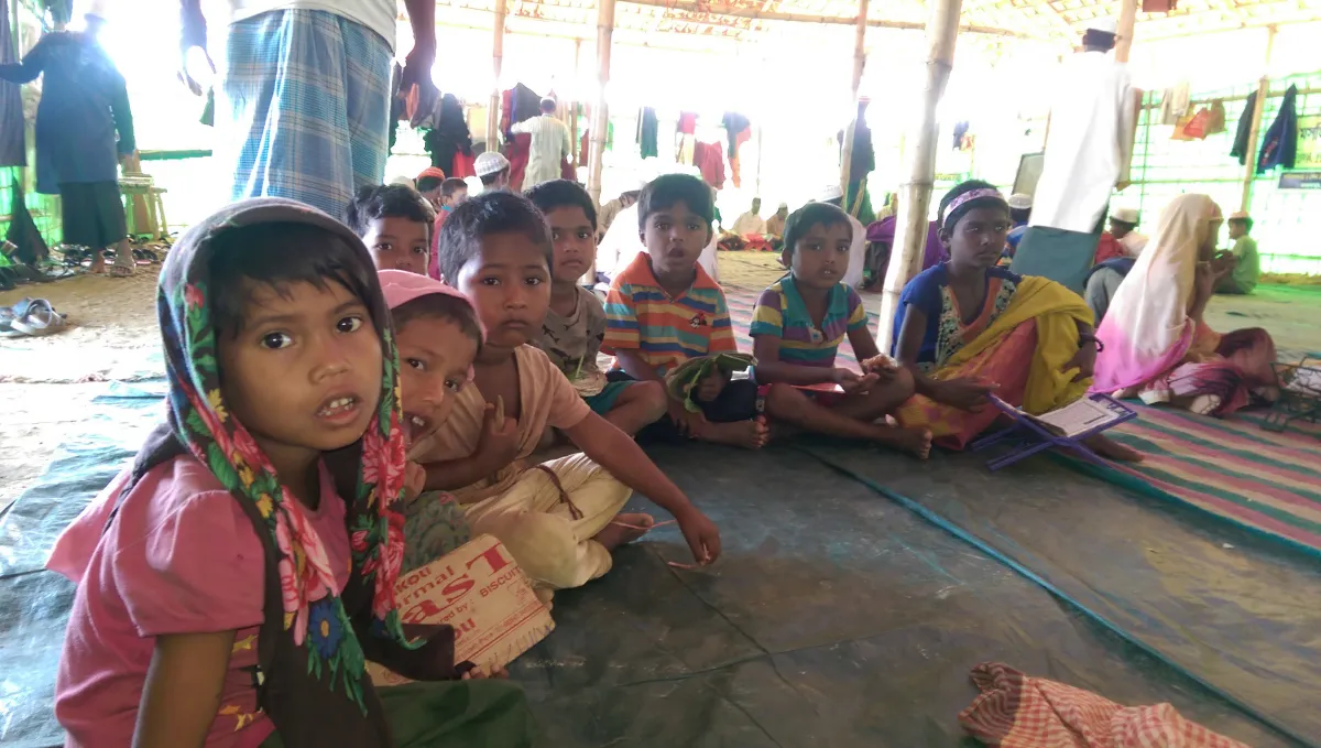 Un groupe d’enfants portant des vêtements colorés sont assis en tailleur sur le sol à l’intérieur d’un abri. Des adultes se tiennent debout derrière eux, et il y a des vêtements accrochés aux murs.
