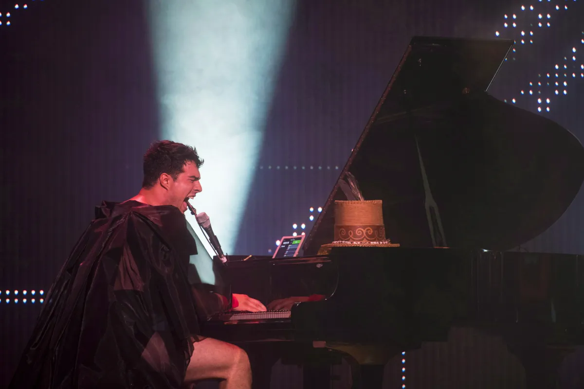 Un homme vêtu d’une cape noire diaphane et d’un short noir joue sur un piano à queue et chante intensément dans un micro. Derrière lui, on voit la lumière d’un projecteur dirigé vers le plafond.