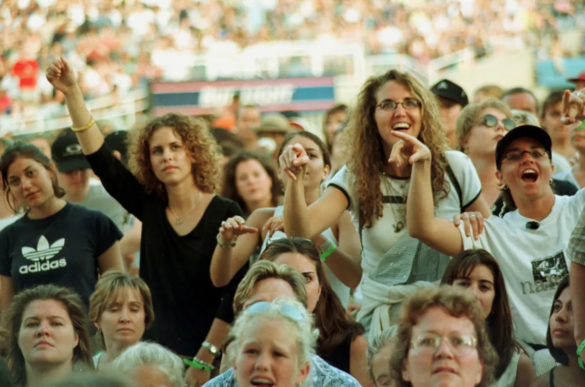 Une foule de femmes, regardant un spectacle hors champ. Un bon nombre d’entre elles sourient, et certaines ont les bras levés. On peut voir une foule plus nombreuse assise dans un stade derrière elles.