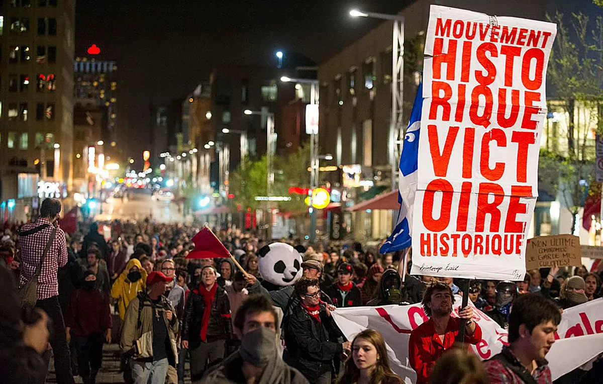 Une mer de jeunes adultes marche dans une rue animée le soir, entourée par des bâtiments et des lampadaires. Une personne tient une pancarte où l’on peut lire « mouvement historique victoire historique ». Une autre porte un costume de panda.