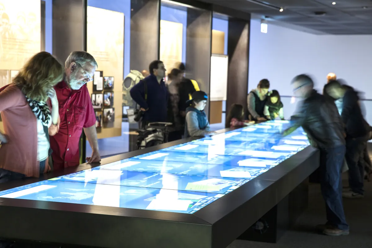 Des membres du public regardent une grande table rectangulaire dont la surface est un écran tactile.
