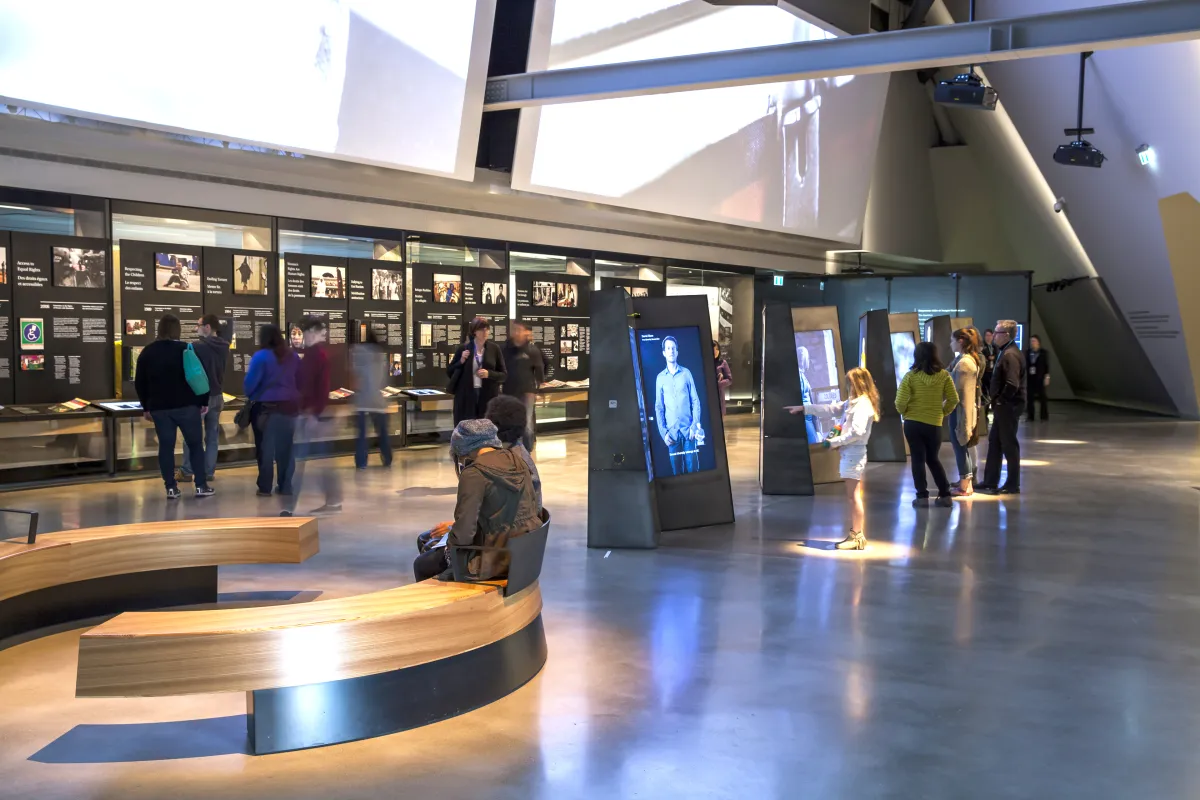 Des gens explorent une galerie contenant des vidéos interactives verticales au centre de la salle et des présentoirs avec texte et images qui longent le mur arrière.