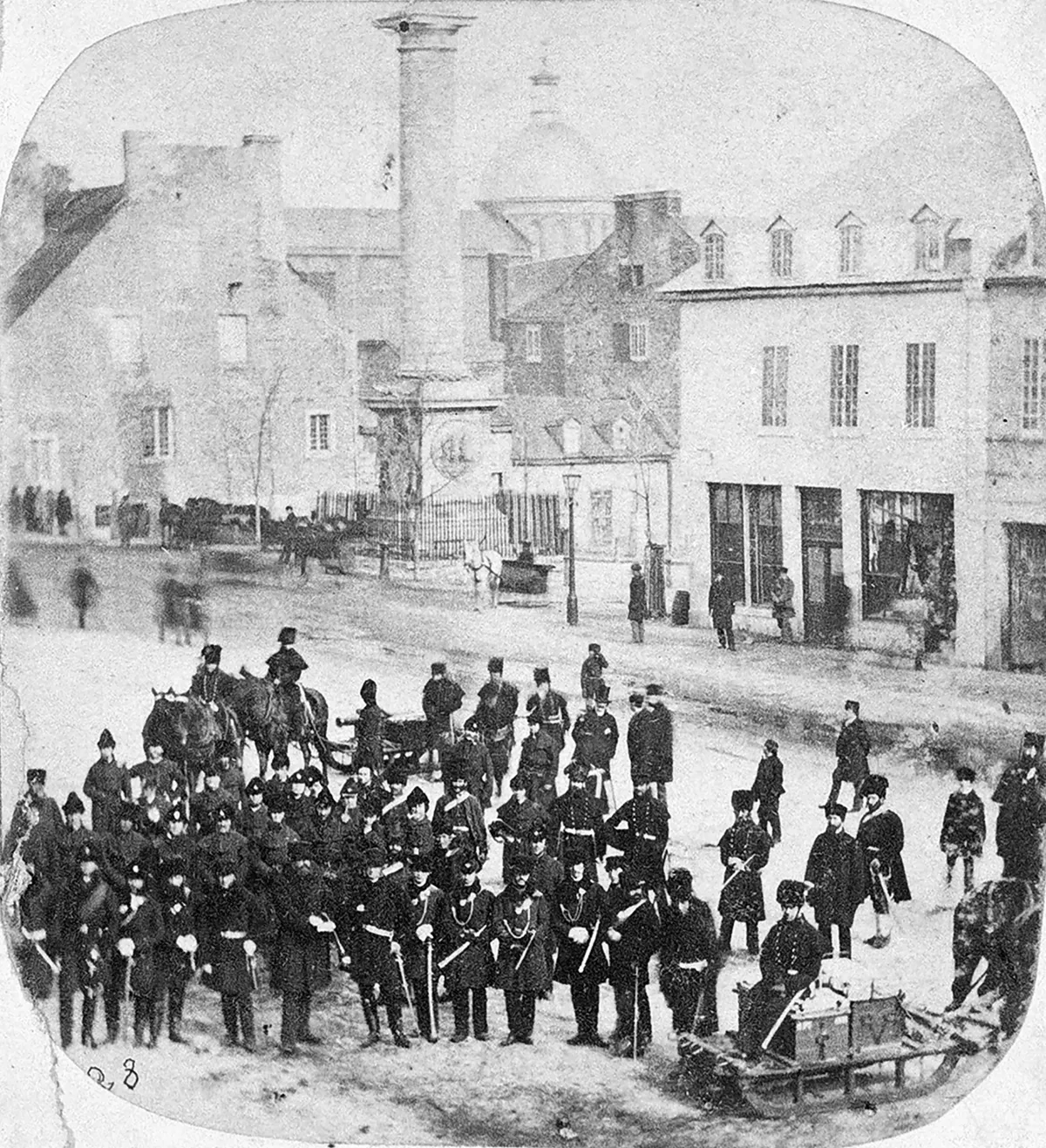 Photo en noir et blanc d’un grand nombre de soldats en uniforme du 19e siècle debout le long d’une rue avec des bâtiments en pierre et en bois.