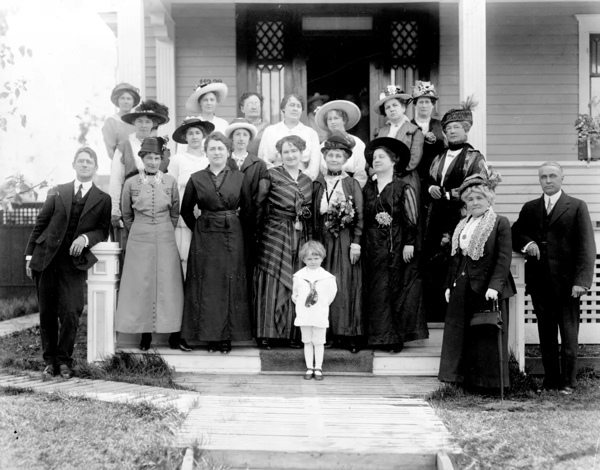Photographie en noir et blanc de 17 femmes, deux hommes et un enfant sur le perron d’une maison. Tous sont vêtus de vêtements du début du 20e siècle.