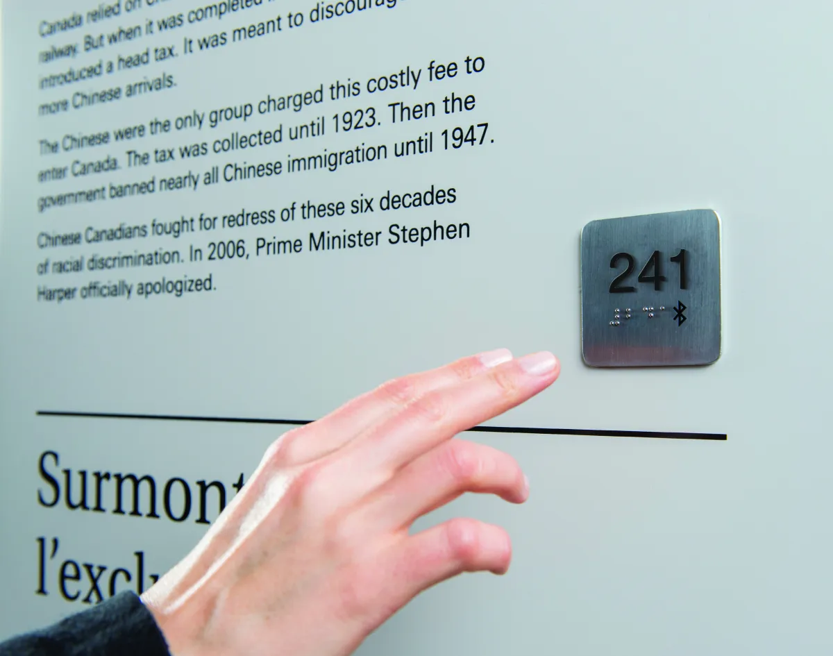 Une main s’avance vers un point d’accès universel, qui est une petite plaque de métal carrée fixée au mur. La plaque porte le numéro 241 en relief et en braille, de même que le symbole Bluetooth.