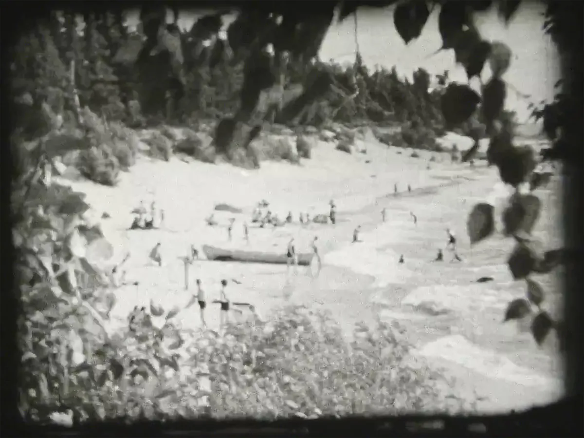 Une photographie en noir et blanc d’une scène de plage, avec des gens sur le sable et dans l’eau, et un canot sur le sable près de l’eau.