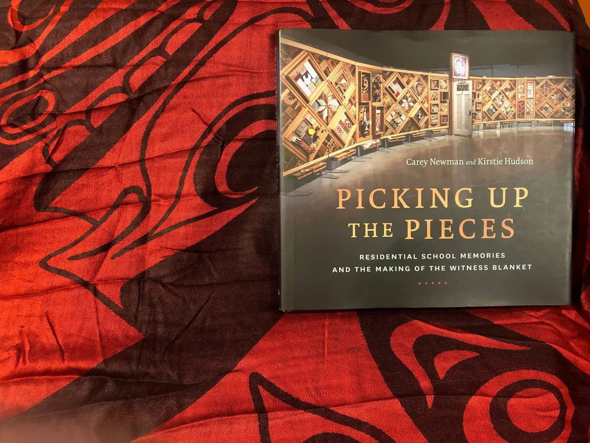 Un livre intitulé « Picking up the Pieces: Residential School Memories and the Making of the Witness Blanket » est posé sur un tissu rouge et noir. La couverture comprend une photographie d’une grande installation artistique à armature de bois.