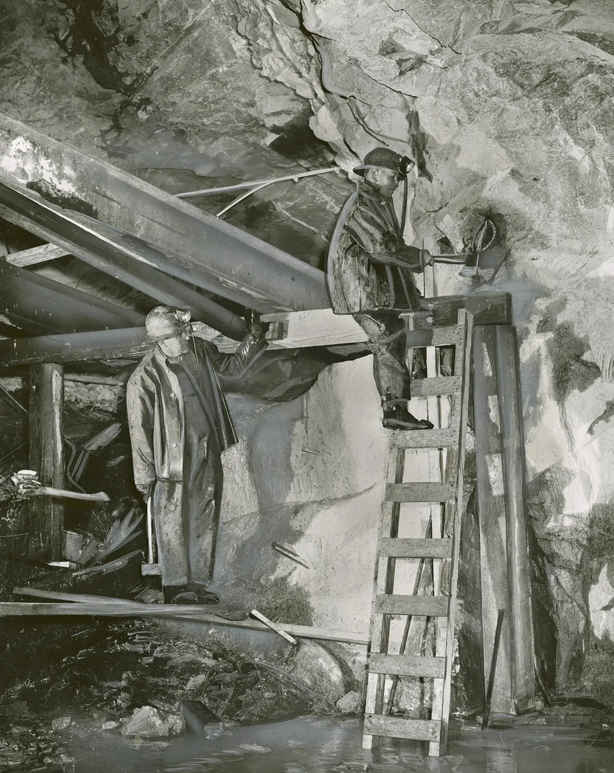 Deux mineurs portant un casque travaillent sous terre dans une mine d'uranium. L'un est debout sur une échelle alors que l'autre le regarde.