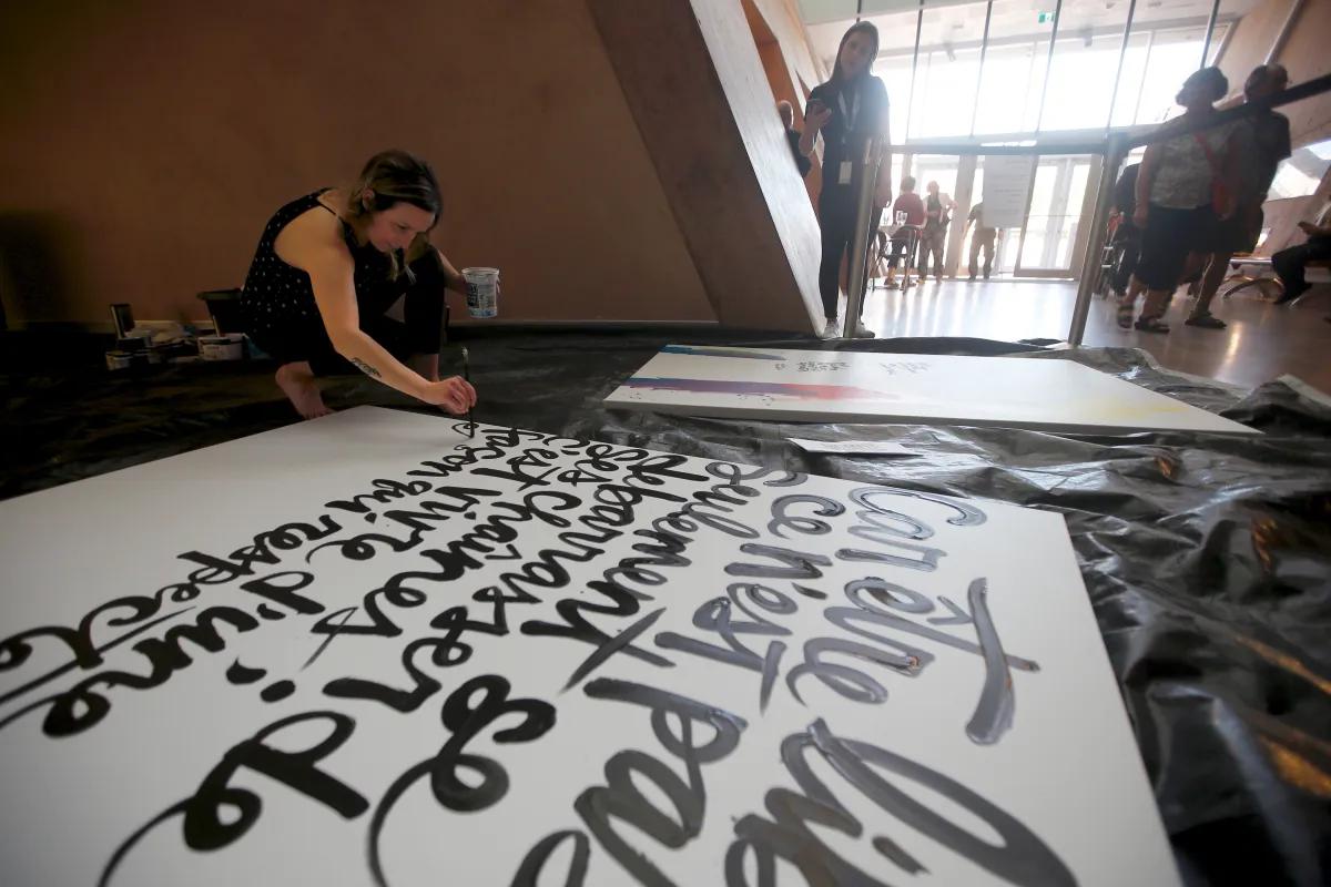 Une femme accroupie sur le plancher peint des caractères d'écriture stylisés sur une grande toile blanche.