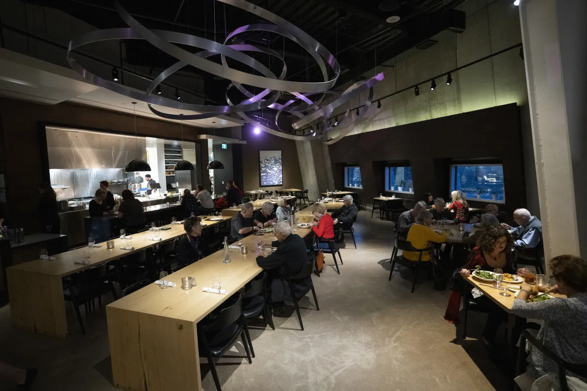 Des personnes assises à des tables et à de longs comptoirs en bois dans un restaurant chic. Une installation artistique en métal est suspendue au plafond.