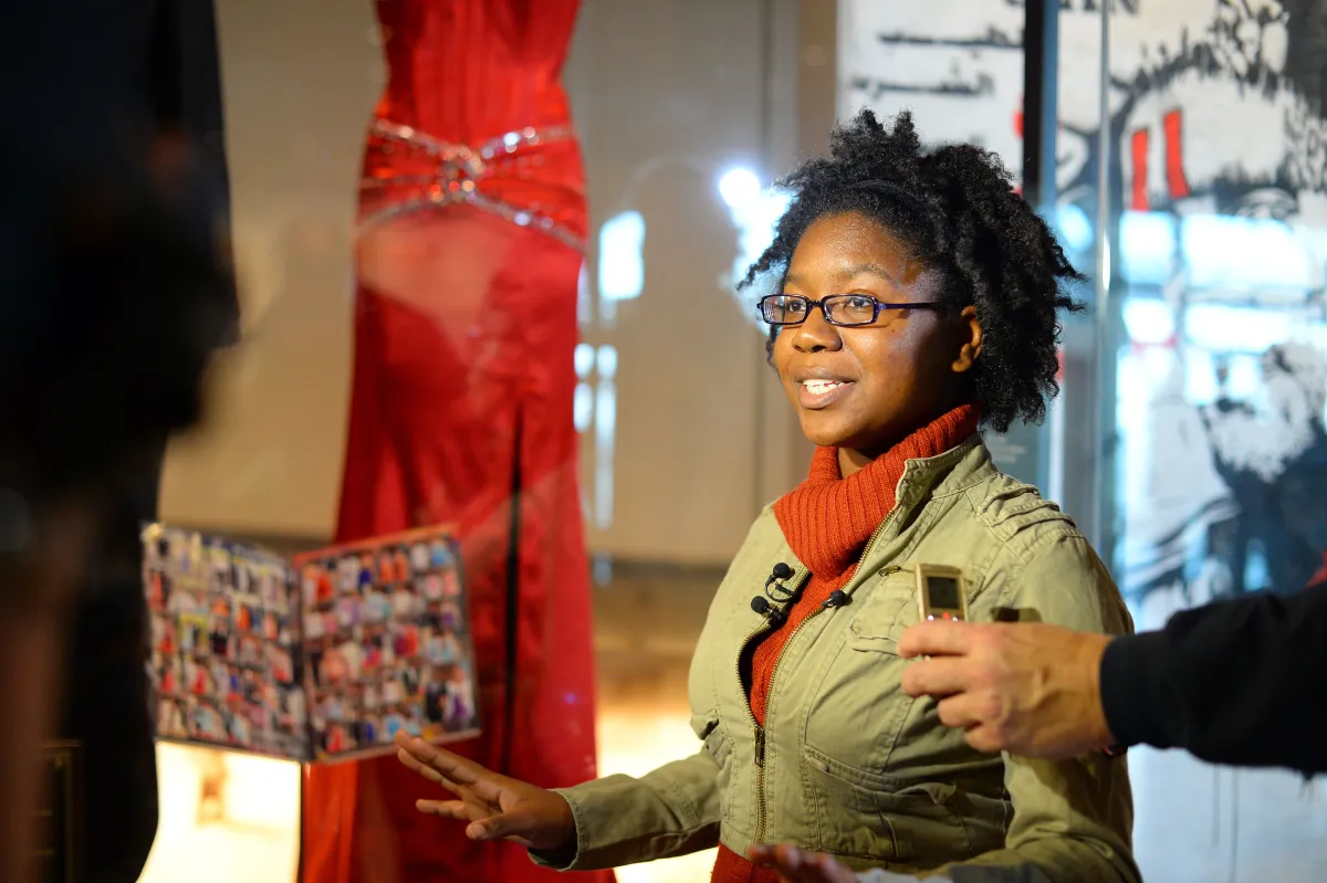 Une jeune femme souriante se tient devant une robe rouge. Elle a un microphone sur le revers de sa veste. Elle parle à quelqu’un qu’on ne voit pas sur la photo.