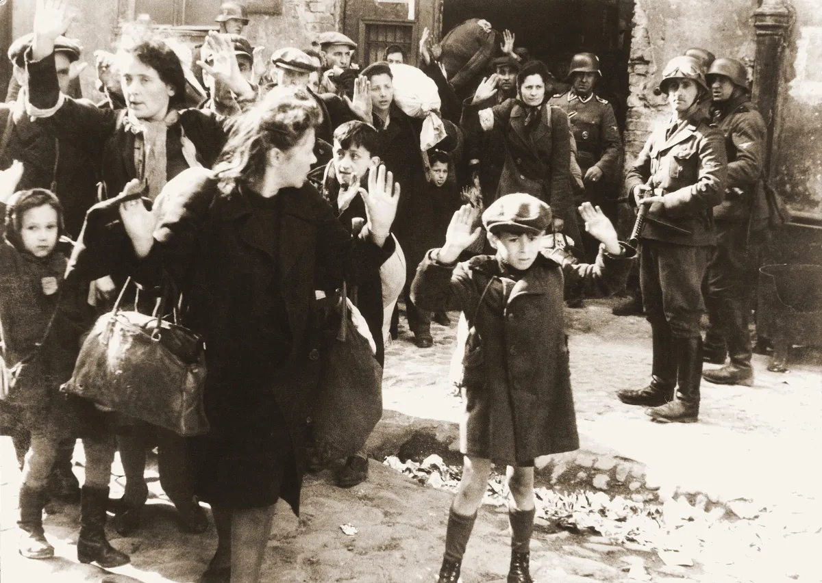Photo en noir et blanc montrant un groupe de personnes les mains levées, sous la menace de soldats armés. Au premier plan, un jeune garçon effrayé.