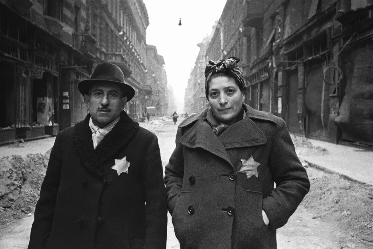 Un homme et une femme vêtus de manteaux arborant une étoile jaune bien visible, dans une rue jonchée de décombres.