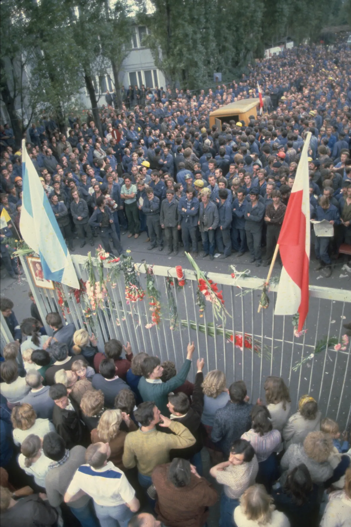 Des milliers de gens en uniforme bleu sont rassemblés d’un côté d’une grande clôture. Des gens en vêtements de tous les jours sont de l’autre côté de la clôture, qui est décorée de fleurs et de deux drapeaux.