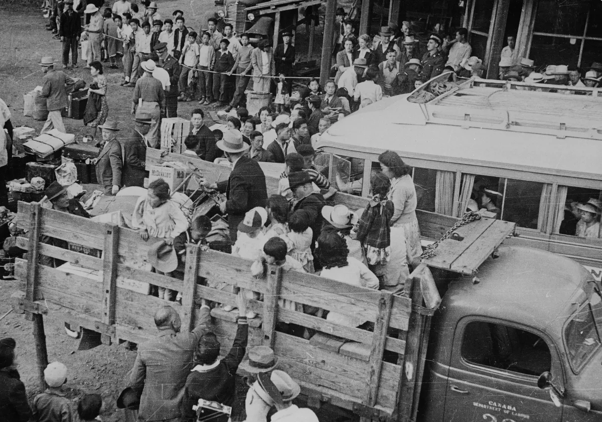 Une photographie en noir et blanc de personnes qui se font embarquer dans des camions alors que d’autres derrière elles font la queue.