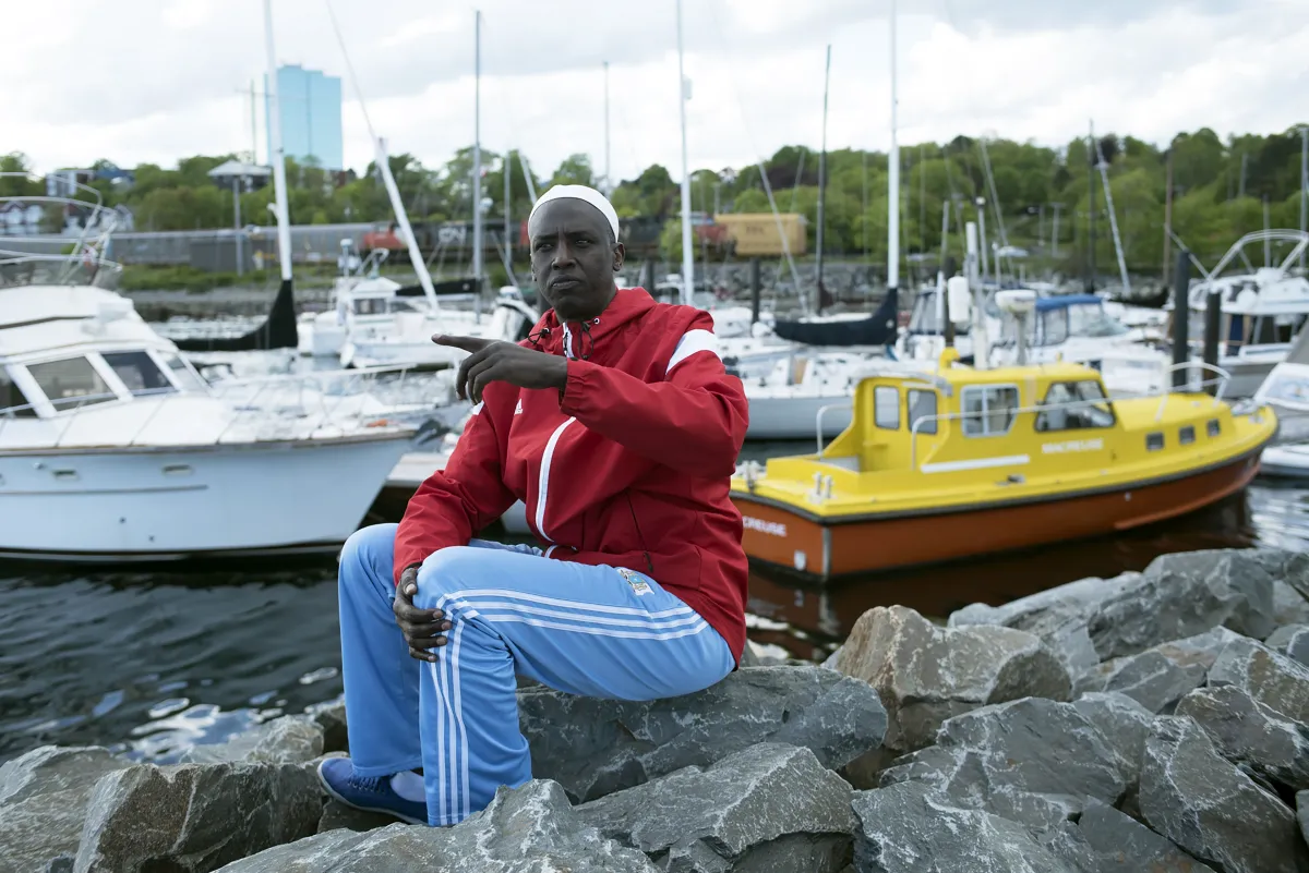 Un homme portant un survêtement de couleurs vives et une calotte blanche est assis sur des rochers, avec des bateaux et un port en arrière-plan.