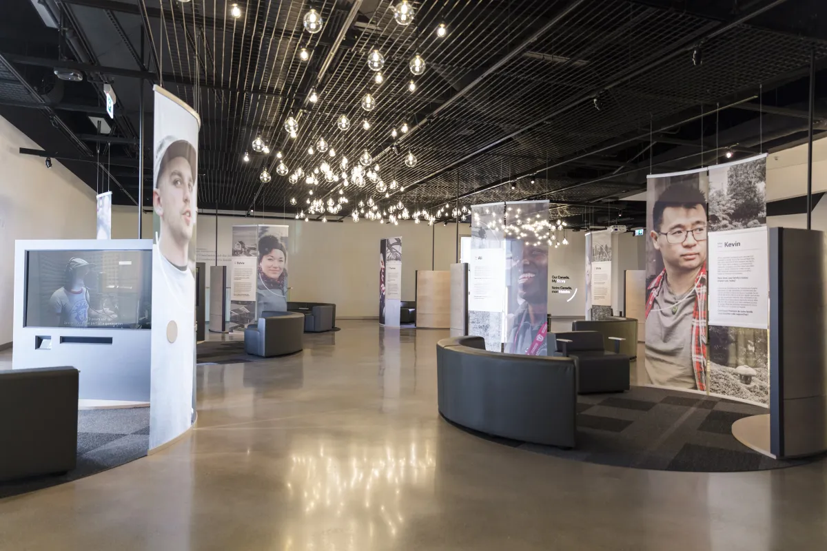 Une exposition muséale comprenant de grands portraits de personnes diverses, des écrans vidéo et des sièges confortables. 