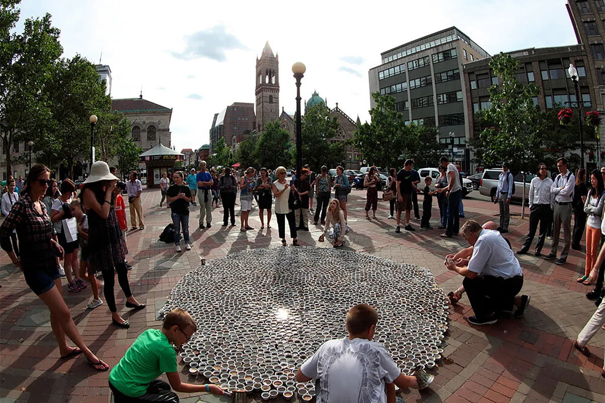 Sur une place de la ville, les gens sont debout ou agenouillés autour d’un grand étalage de tasses à café disposées sur le sol.