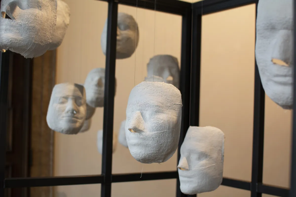 Plusieurs visages en plâtre blanc sont suspendus à des cordes, à l’intérieur d’une cage.