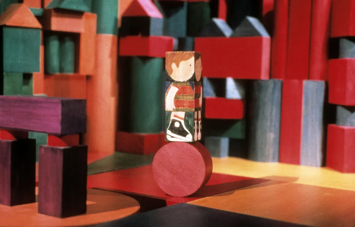 Un bloc de bois, sur lequel est peinte l'image d'un enfant, est posé sur un bloc rond. Des blocs de formes, de couleurs et de tailles différentes apparaissent à l'arrière-plan.