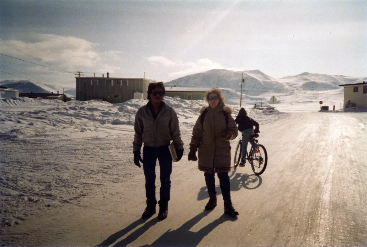 Deux personnes portant un manteau d’hiver se tiennent dans un paysage enneigé, devant des collines et des bâtiments en arrière-plan.
