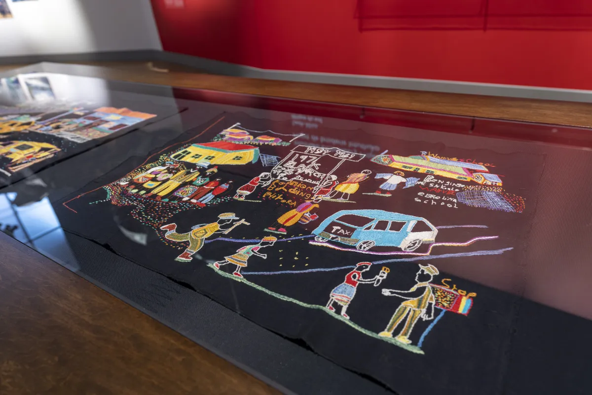 Un morceau de broderie coloré sur un fond noir représente une scène avec des personnes, des maisons et des véhicules.