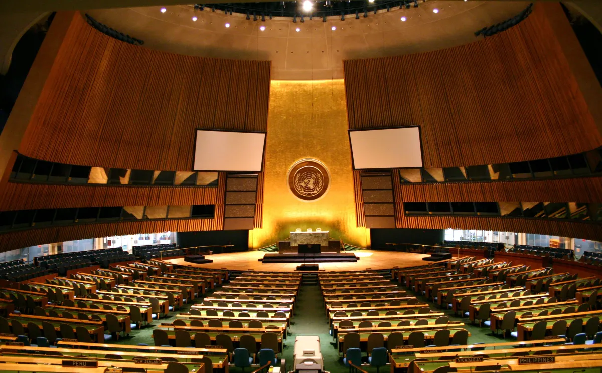 Une grande salle avec des rangées de bureaux et de chaises faisant face à un haut mur orné du logo de l’ONU.