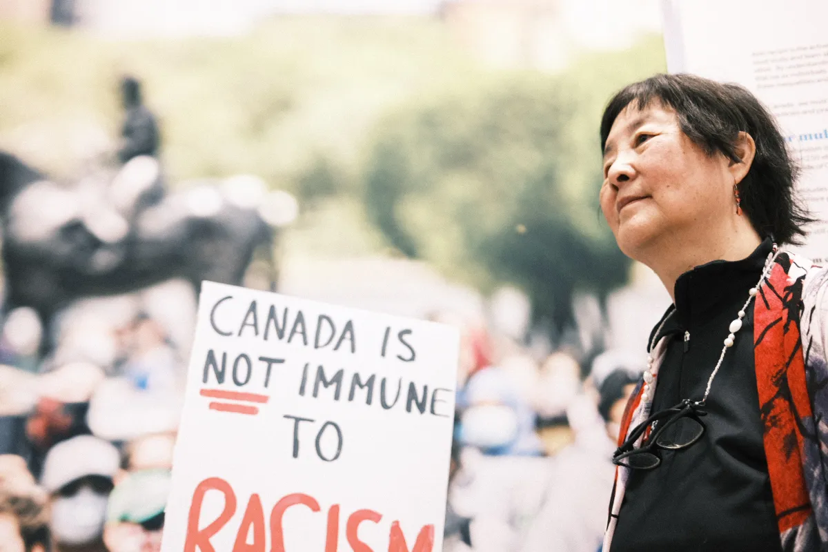 Une femme se tient à côté d’une grande photo d’une foule où quelqu’un tient une pancarte sur laquelle on peut lire en anglais: « Le Canada n’est pas à l’abri du racisme ».