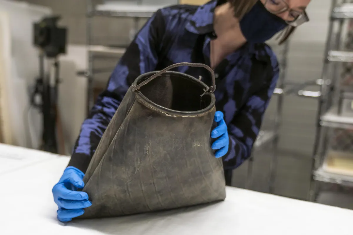 Une femme tient un sac en caoutchouc noir éraflé avec une poignée métallique courbée de forme grossière sur le dessus.