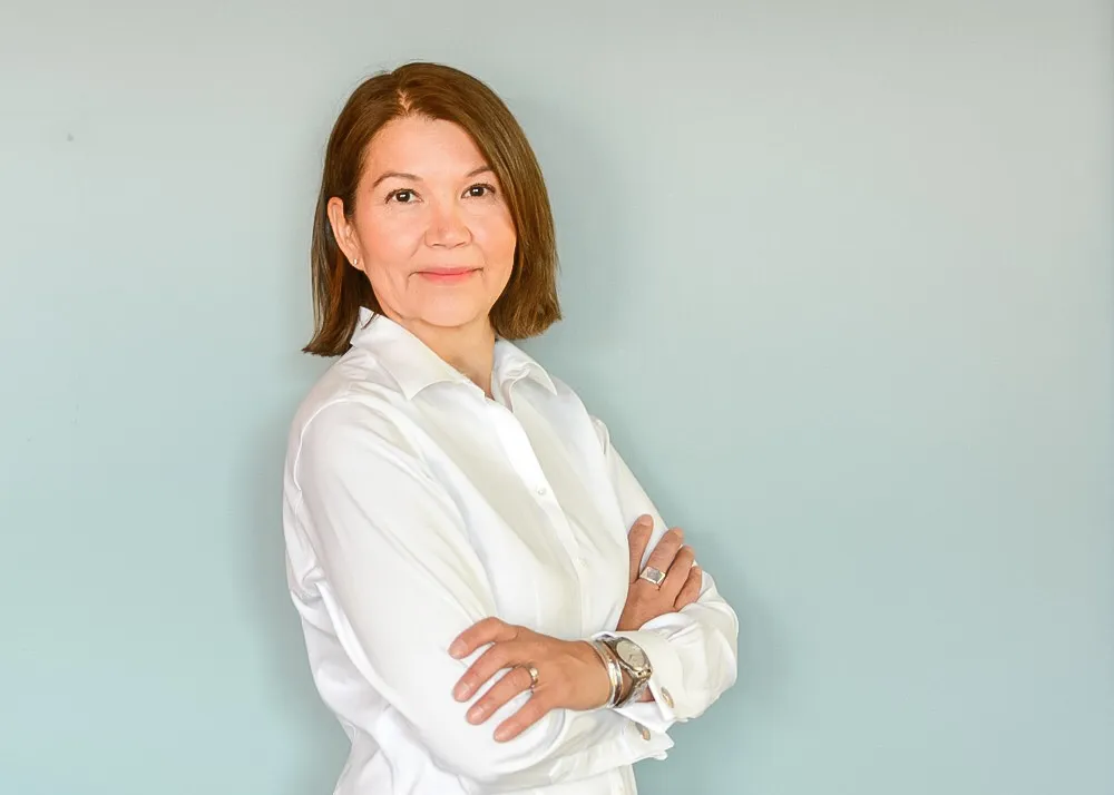 Une femme avec des cheveux bruns raides et longs aux épaules et un chemisier blanc.