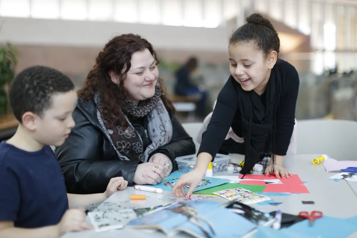 Un parent souriant et deux enfants sont assis à une table, avec des matériaux de bricolage comprenant des marqueurs, des ciseaux, de la colle et du papier de construction disposés devant eux.