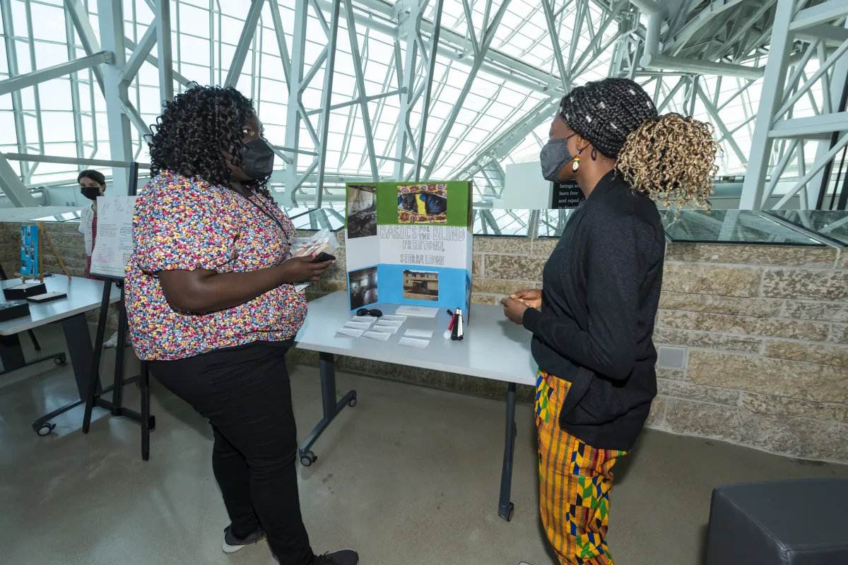 Deux femmes parlant entre une affiche qui dit "Basics for the blind, Freetown, Sierra Leone".