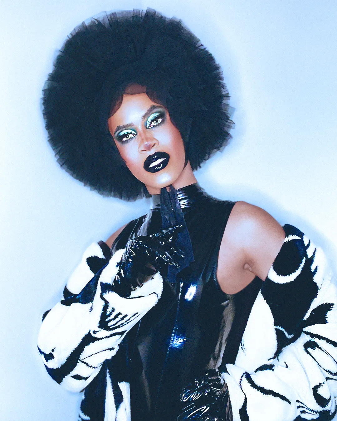 Un homme biracial habillé en drag. Il porte une coiffe noire, des gants en cuir noir, une robe en cuir noir, un pull noir et blanc.