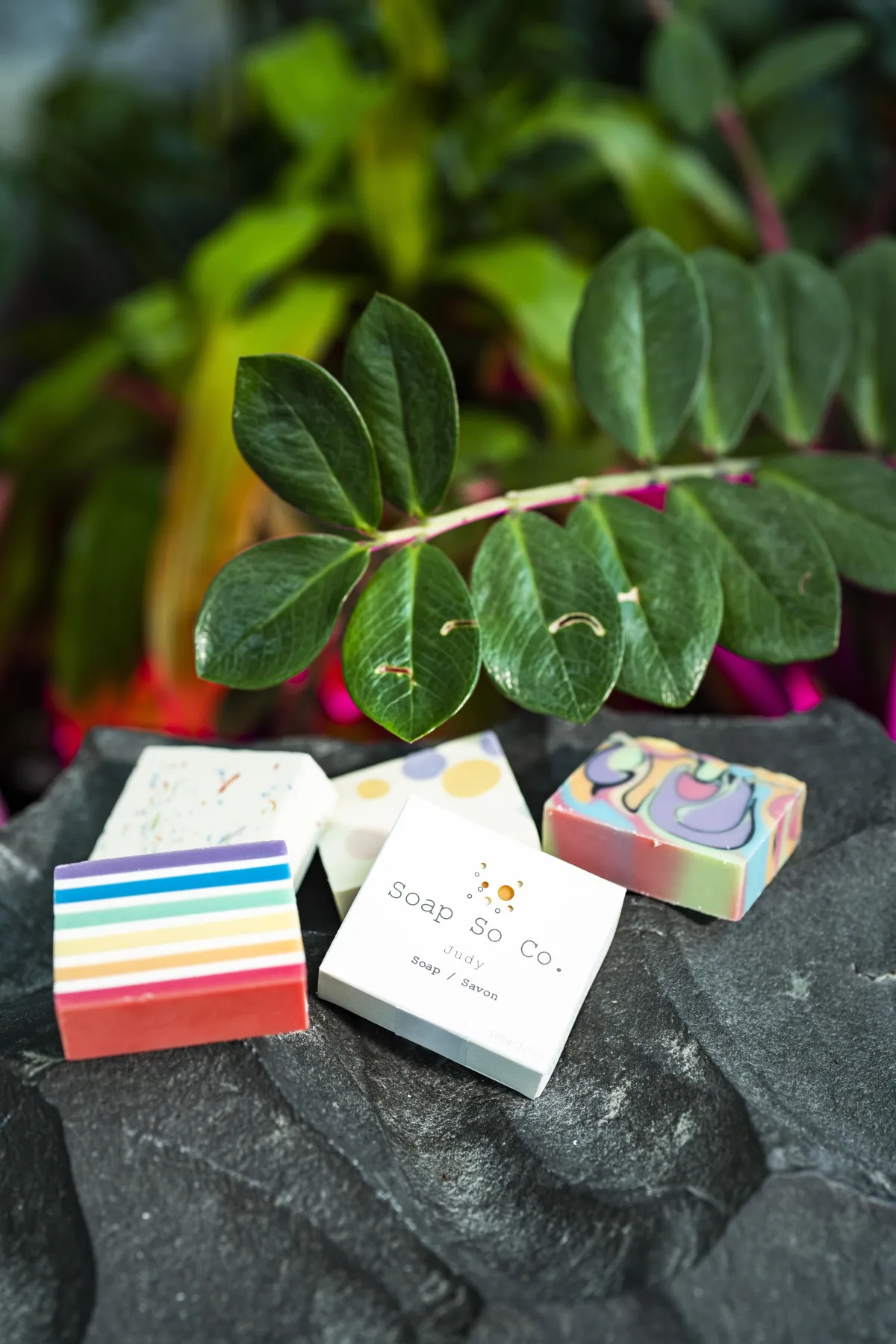 Un pain de savon avec des bandes colorées ressemblant au drapeau de la Fierté se trouve au premier plan, à côté d'une boîte sur laquelle on peut lire "Soap So Co". À l'arrière-plan se trouvent d'autres pains de savon aux couleurs et aux motifs variés.