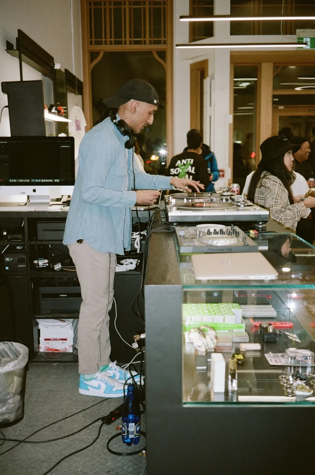 Suraj, un DJ portant une casquette de baseball à l’envers, fait tourner des disques sur un comptoir en verre lors d’un événement.