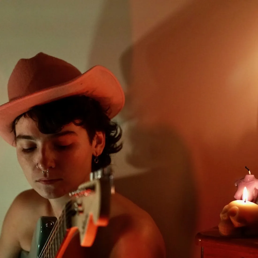 Une musicienne joue de la guitare. Ses cheveux foncés bouclent sous un chapeau de cowboy marron en feutre; ses épaules sont nues, et son nez et ses oreilles sont percés. Sur le côté, une bougie en forme de crâne brûle à côté d’une bougie rose éteinte en forme de torse de femme.