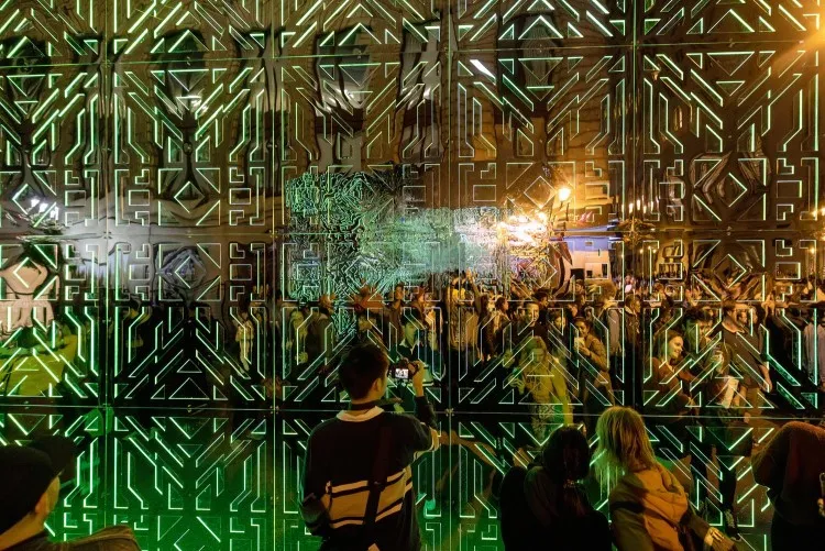 Cinq personnes se tiennent devant des miroirs joints aux motifs complexes qui reflètent des lumières vertes et une foule.