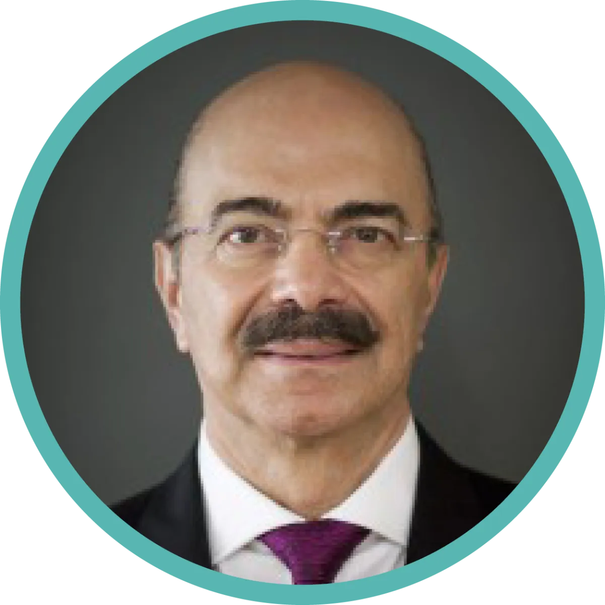Un homme à la moustache sombre et aux petites lunettes sans monture porte un complet sombre et une cravate violette.