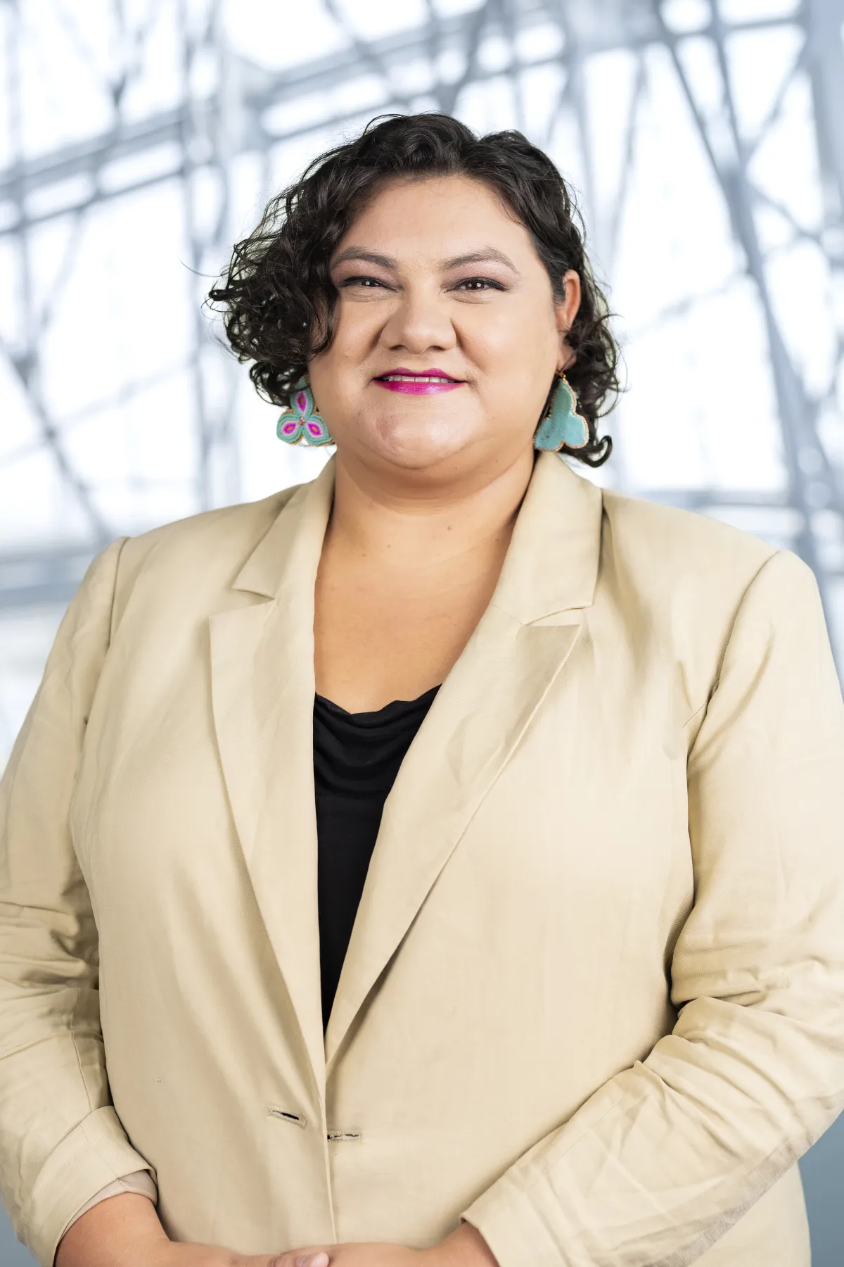 Une femme autochtone aux cheveux bruns courts porte un blazer beige, superposé à un chemisier noir. Elle porte des boucles d’oreilles en perles roses, vertes et blanches. Elle est de face et sourit à l’appareil photo.