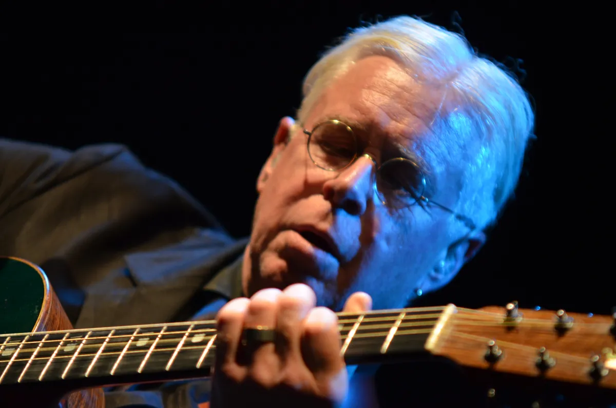 Gros plan d’un homme à lunettes et aux cheveux blancs jouant de la guitare.