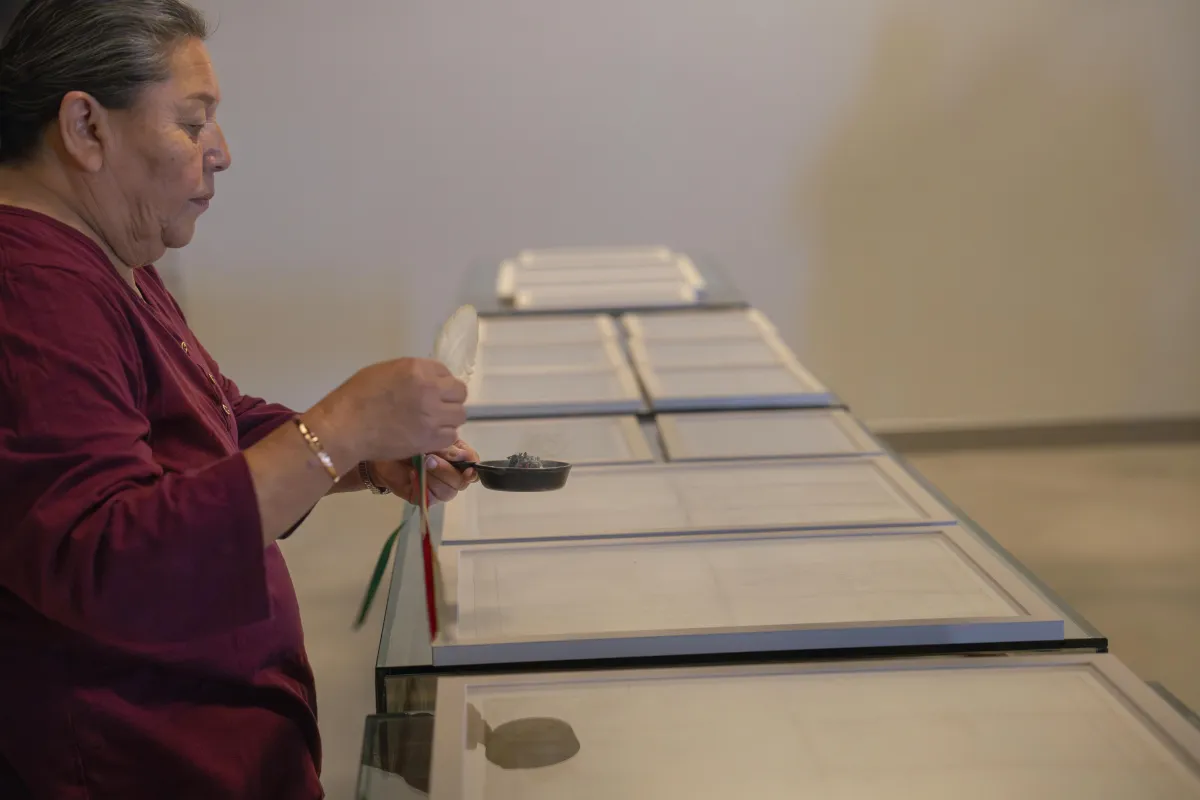 Des documents encadrés sont posés sur une table tandis que l'aînée Hazel Copenace les barbouille, tenant un bol à fumée dans sa main gauche et une plume d'aigle dans sa main droite.