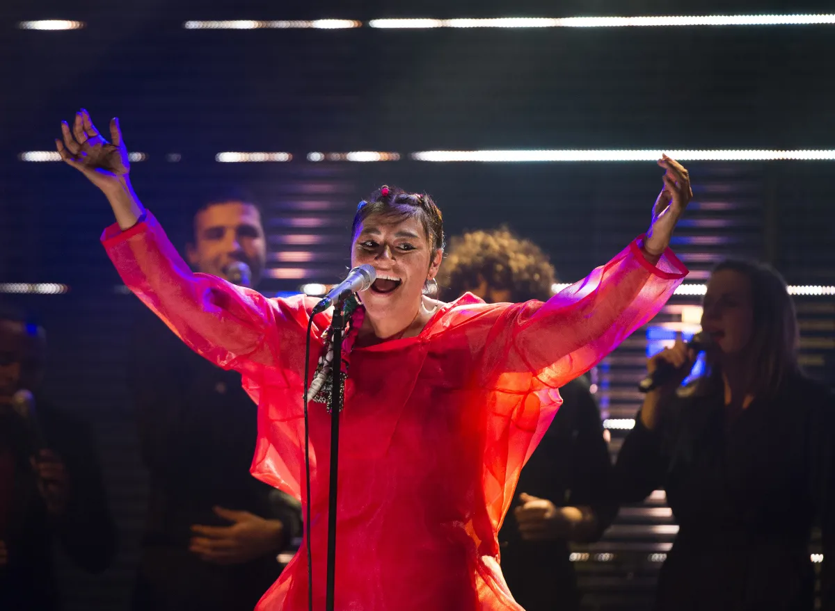  Une femme chante à un micro, en faisant de grands gestes avec ses bras. Elle porte une robe rouge foncé, diaphane, avec de longues manches.