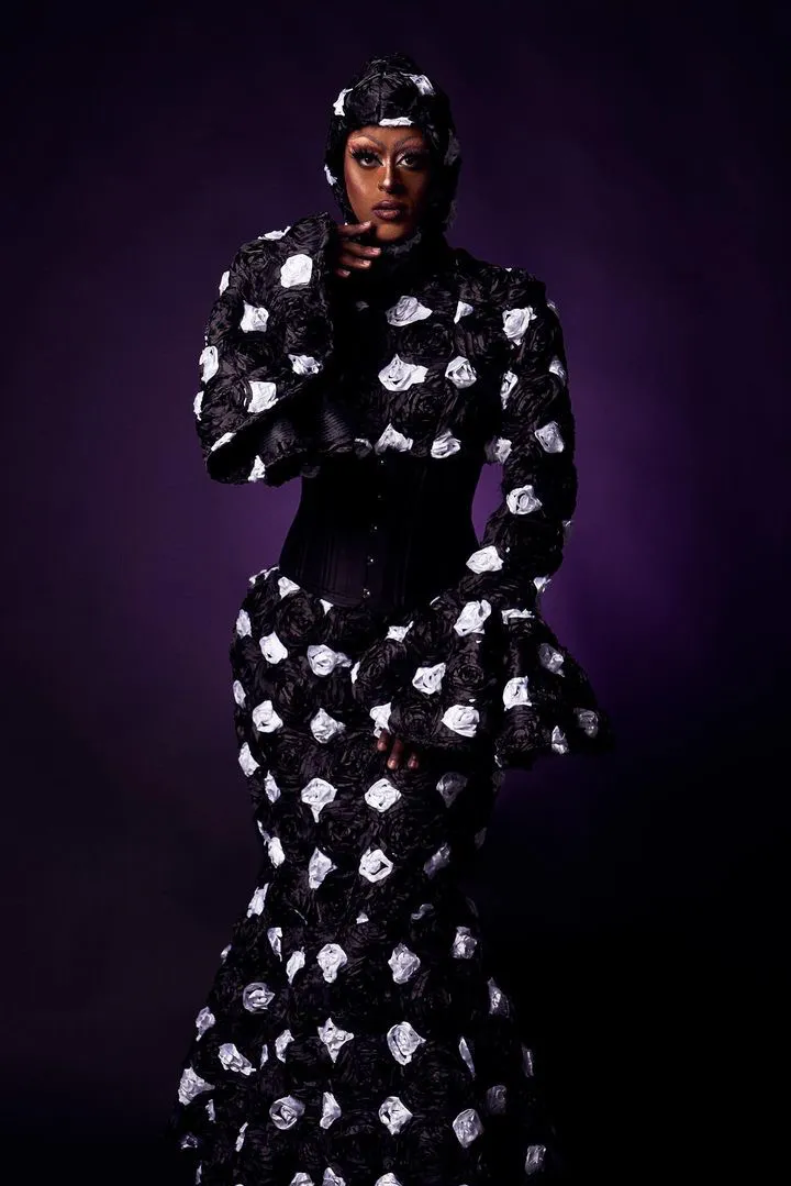 Une drag queen vêtue de la tête aux pieds d'une longue robe noire de style sirène ornée d'un motif imprimé blanc.