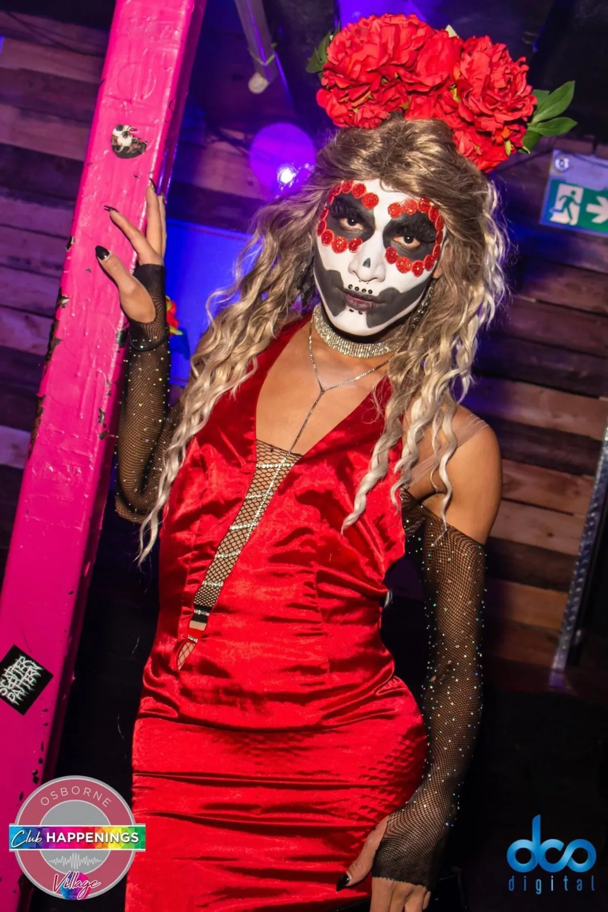 Une drag queen aux longs cheveux blonds ondulés surmontés de grandes roses rouges, au visage peint et à la robe de satin rouge.