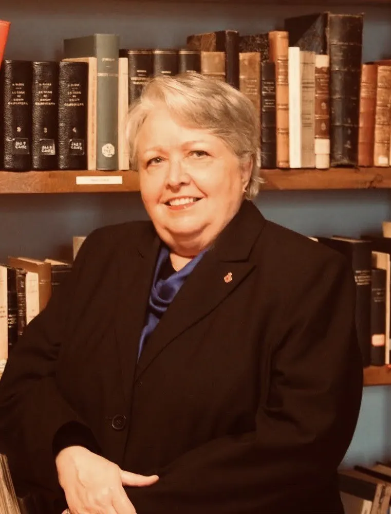 Randi Gage est une femme d’un certain âge, aux cheveux courts et pâles, vêtue d’un blazer noir. Elle est assise devant une bibliothèque.