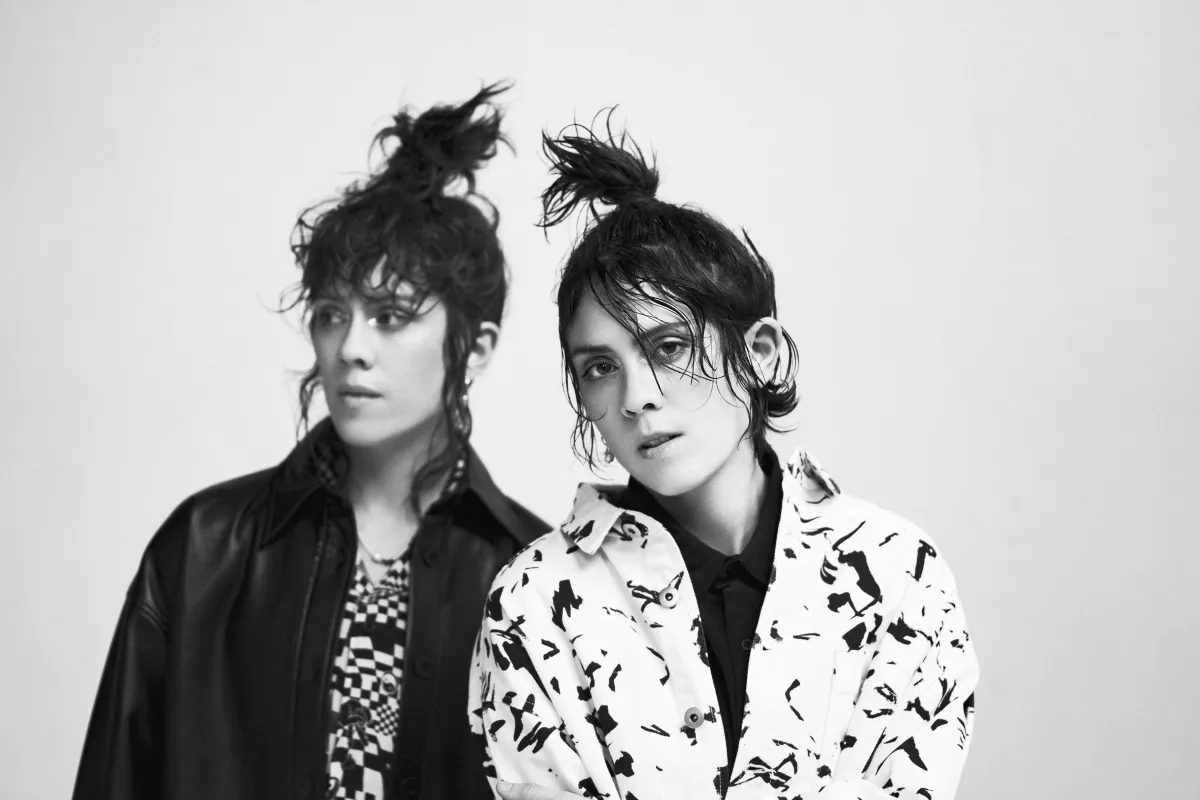Deux jeunes personnes avec les cheveux noués en désordre. L’une porte une veste en cuir noir, l’autre une veste blanche avec un motif de taches noires.