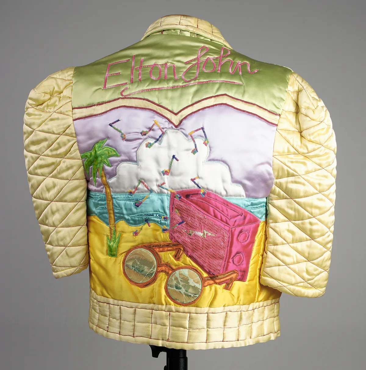 Le dos d’une veste en satin matelassé jaune brillant, décoré des mots « Elton John » au-dessus d’une scène de plage avec un ciel violet, un gros nuage blanc, une mer bleue, du sable jaune foncé, un palmier, une radio rose foncé d’où s’échappent des notes de musique, et une paire de lunettes de soleil rondes au premier plan.