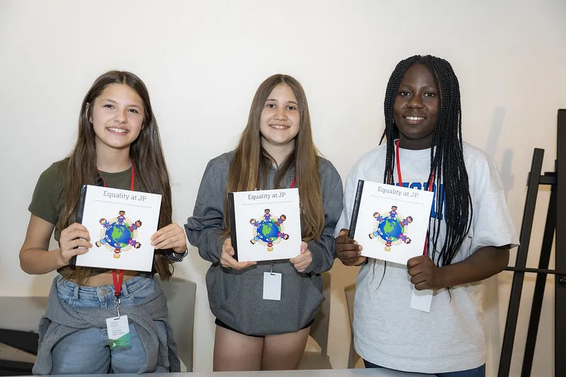 Trois élèves tiennent des exemplaires d’un livre qu’elles ont créé. La couverture du livre porte un titre anglais qui dit « L’égalité à JP » et présente un dessin de la terre avec des élèves tout autour.