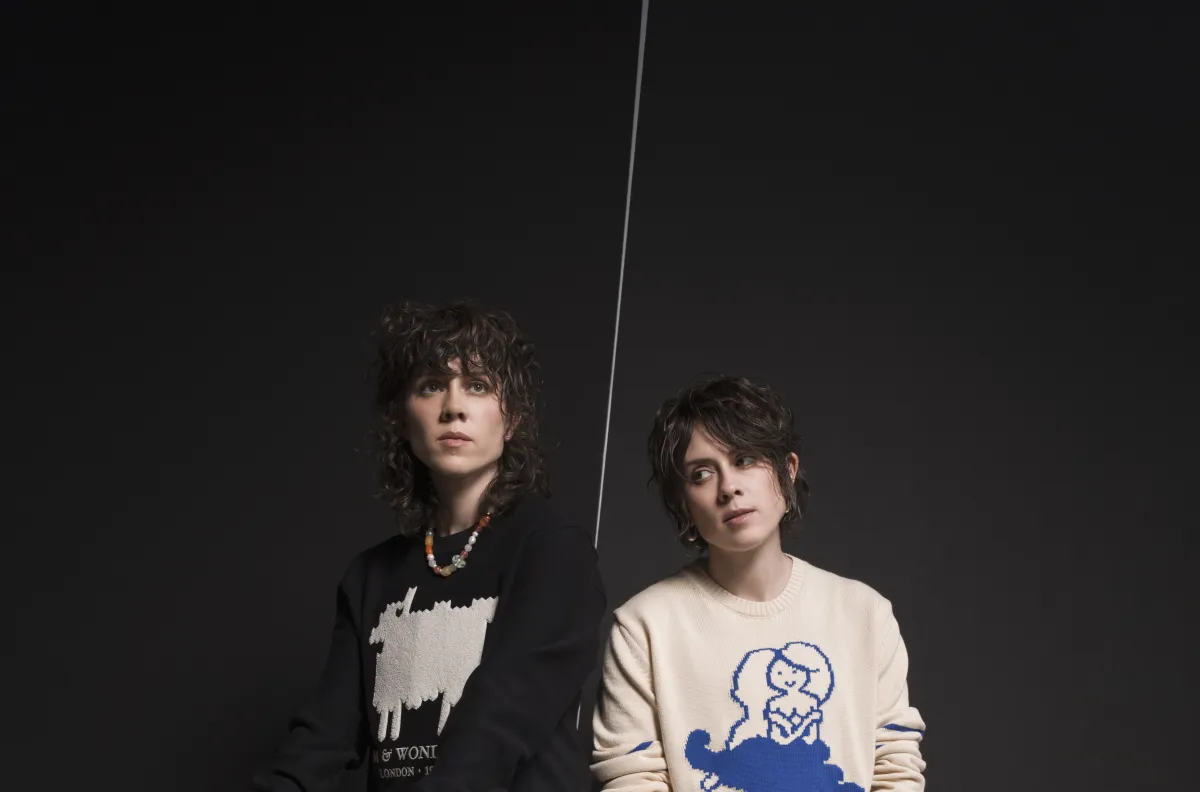Deux personnes aux cheveux mi-longs posent sur un fond noir. L'une porte un pull noir avec un mouton blanc sur le devant, l'autre porte un pull blanc avec une sirène bleue.